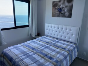 Quarto com vista para o oceano, cabeceira branca com detalhes em tufos e colcha xadrez.