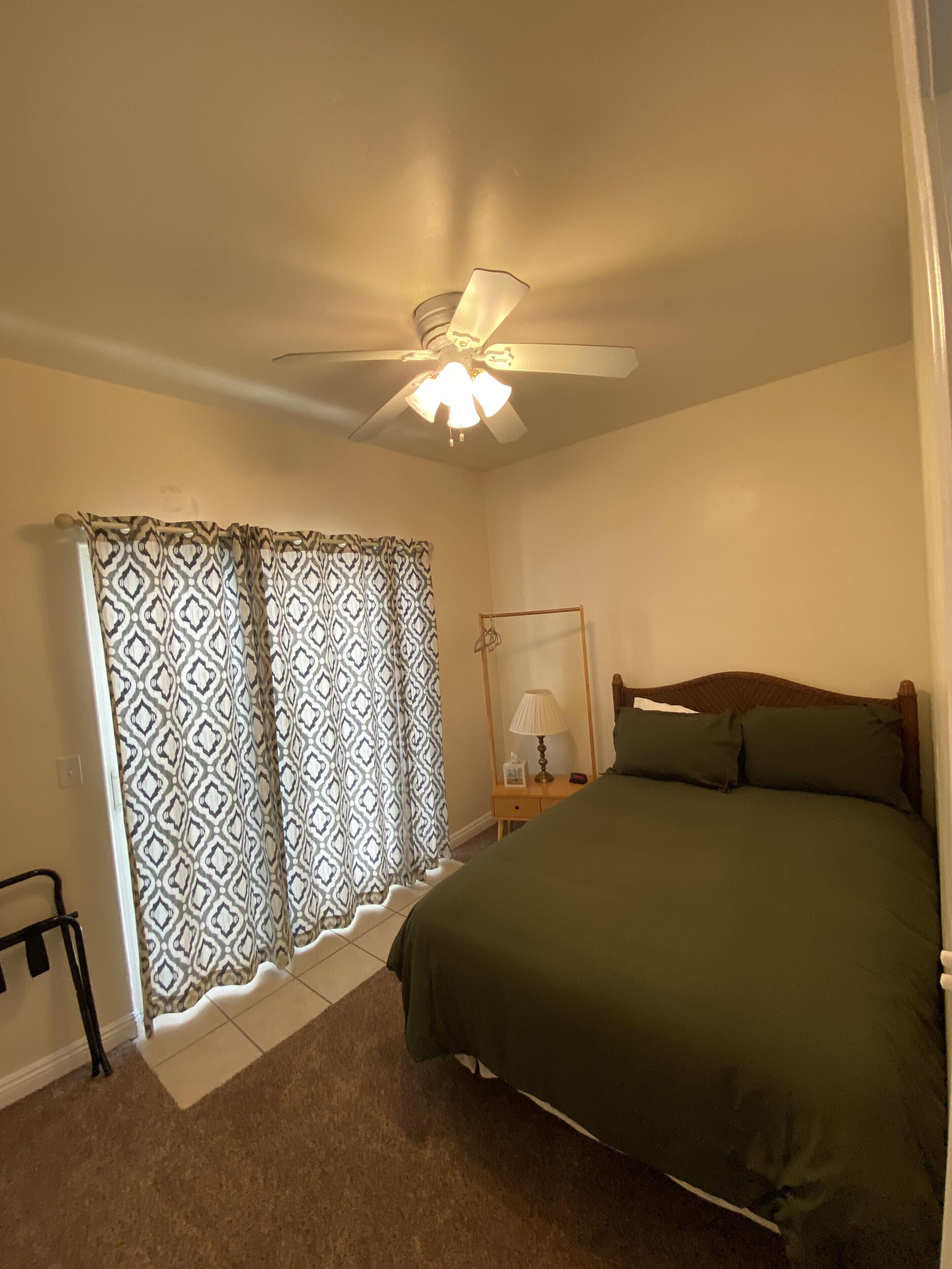 A bedroom with a bed, a nightstand, a lamp, a mirror, a ceiling fan, and patterned curtains.