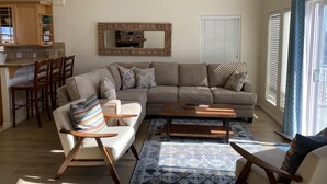 Ein modernes Wohnzimmer mit einem Ecksofa, zwei Sessel, einem Couchtisch und einem gemusterten Teppich. Es gibt eine Küchenzeile mit Holzschränken sowie einen Esstisch mit Stühlen. An der Wand hängt ein großer Spiegel, und ein Fenster mit Jalousien ist sichtbar.