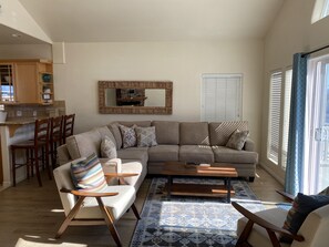 A modern living room with a sectional sofa, two armchairs, a coffee table, and a patterned rug. There is a kitchen area with wooden cabinets and a dining table with chairs. A large mirror hangs on the wall, and a window with blinds is visible.