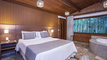 A modern hotel room with a large bed, a wooden headboard, a built-in bench, a large window with a forest view, and a marble bathtub.