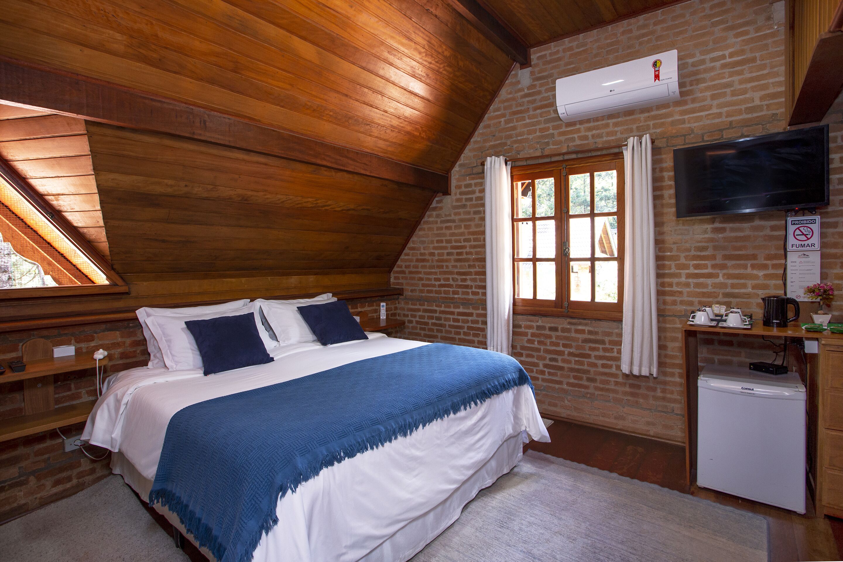 A bedroom with a wooden ceiling, a brick wall, a bed with white and blue bedding, a small refrigerator, a TV, and a window with curtains.