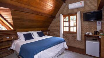 A bedroom with a wooden ceiling, a brick wall, a bed with white and blue bedding, a small refrigerator, a TV, and a window with curtains.