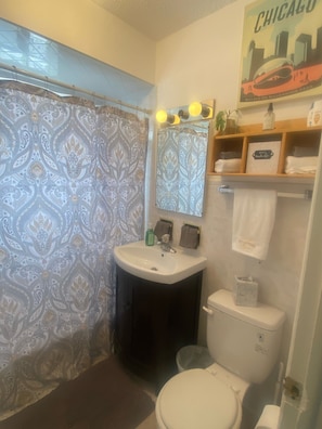 A bathroom with a patterned shower curtain, a sink with a mirror above it, a toilet, and a shelf with towels and toiletries. - Lovely 2 bedrooms apartment 10min from O'HARE CHICAGO (Rosemont)