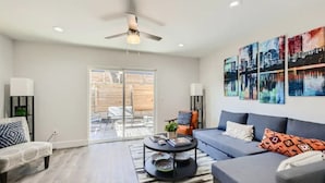 A modern living room with a grey sofa, a coffee table, and a ceiling fan. There are two wall-mounted lamps and a large window with a view of a wooden deck.