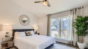 A hotel room with a bed, nightstands, a ceiling fan, a large window with curtains, and a potted plant.