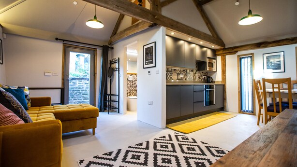 A modern living room with a yellow sofa, a wooden dining table, and a kitchen area with grey cabinets and a yellow rug.
