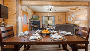 Une salle à manger en bois avec une table dressée pour quatre personnes, une cheminée et un téléviseur en arrière-plan.