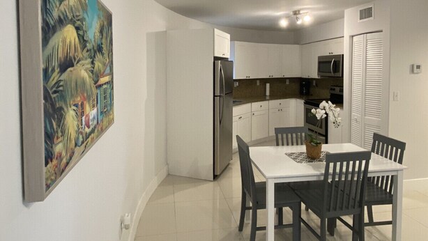 Uma cozinha moderna com armários brancos, uma geladeira de aço inoxidável e uma área de jantar com mesa e cadeiras brancas.