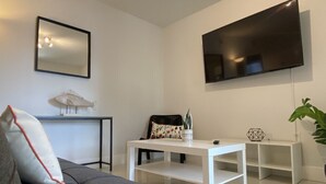 Uma sala de estar moderna com um sofá cinzento, uma mesa de centro branca, uma TV de tela plana na parede, uma mesinha lateral com um objeto decorativo e um vaso de planta.