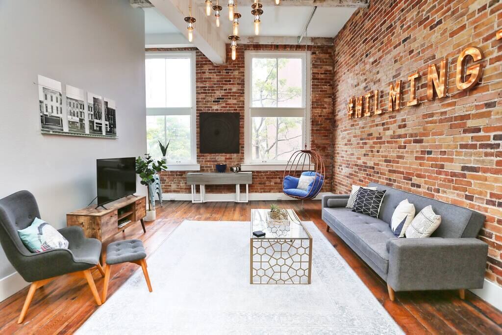 A modern living room with a brick wall, a grey sofa, a black armchair, a glass coffee table, and a hanging light fixture.