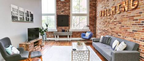 A modern living room with a brick wall, a grey sofa, a black armchair, a glass coffee table, and a hanging light fixture.