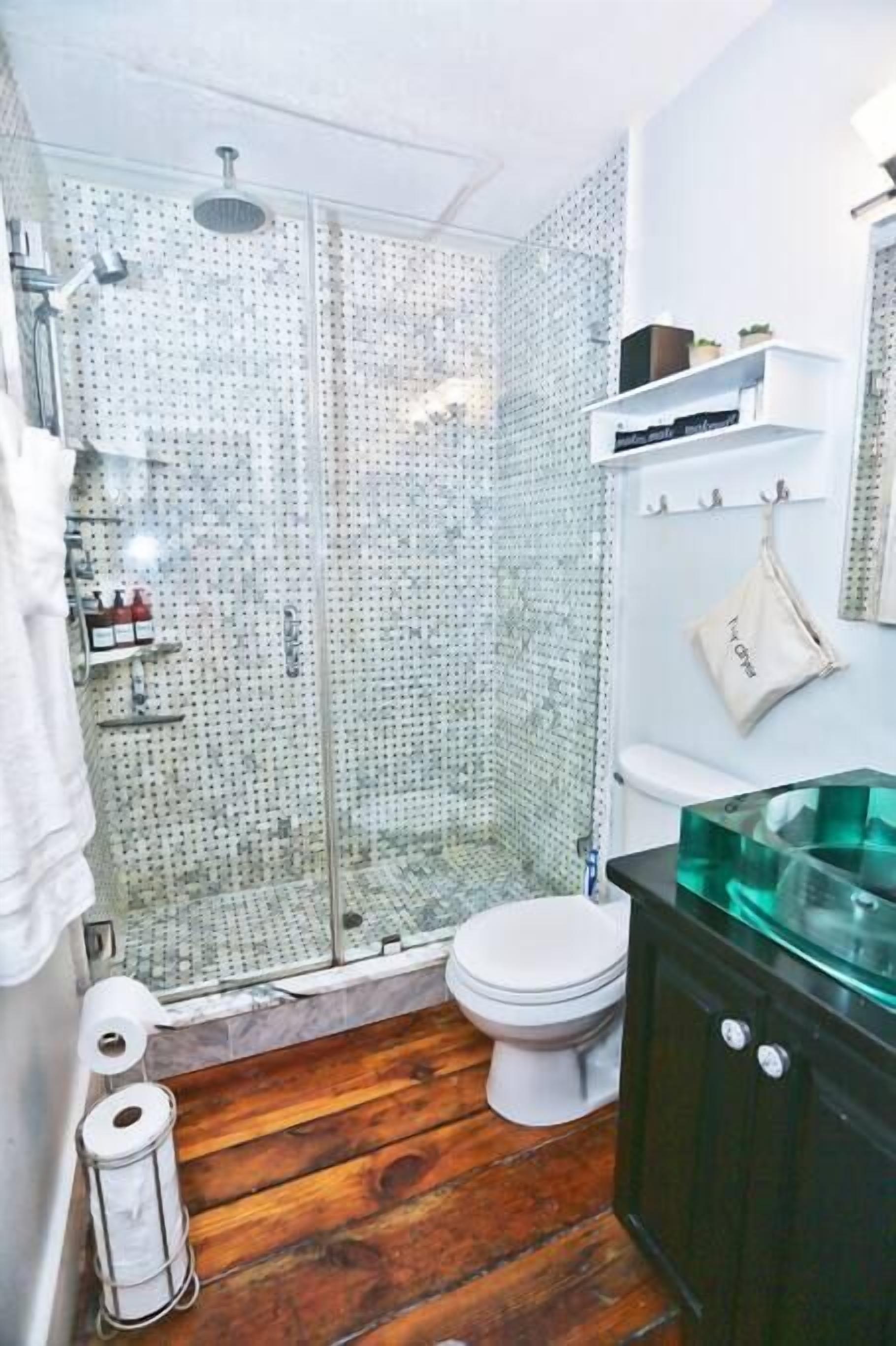 Un baño con cabina de ducha de vidrio, inodoro blanco, piso de madera y un mueble negro con lavamanos de vidrio.