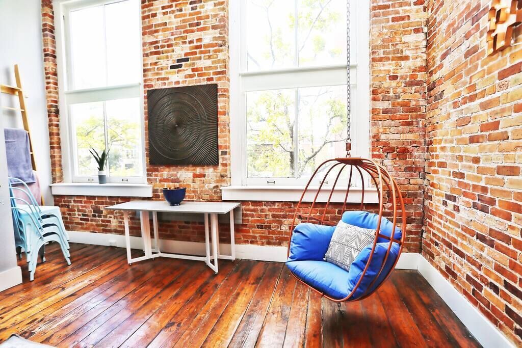 A room with exposed brick walls, a hanging chair with blue cushions, a white table, and a window with a view of trees.