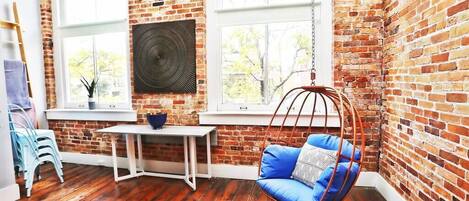 A room with exposed brick walls, a hanging chair with blue cushions, a white table, and a window with a view of trees.
