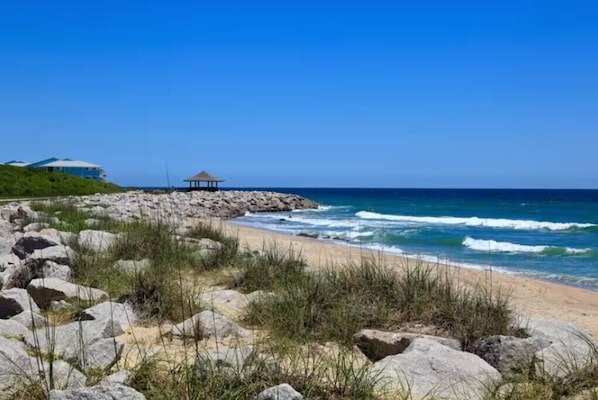 A beach with a rocky shoreline, a thatched structure, and a building in the background.