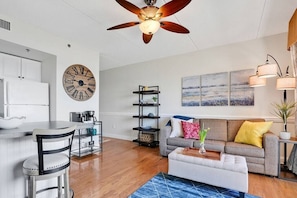 A modern living room with a wooden ceiling fan, a round wall clock, a grey sofa with yellow cushions, a white coffee table, and a blue rug.