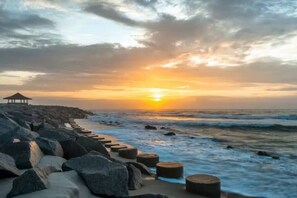 A coastal scene with a wooden pier extending into the sea, large rocks lining the shore, and the sun setting over the horizon. - ⭐️ ⭐️ ⭐️ ⭐️ ⭐️ RIVER FRONT! Battleship View! (Wilmington)
