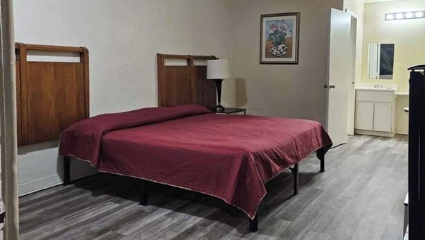 A bedroom with a bed, wooden headboard, a nightstand, a lamp, a painting, and a bathroom with a sink and mirror. - Pharr Executive Inn (Pharr)