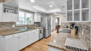 Una cocina moderna con armarios blancos, encimeras de granito, un refrigerador de acero inoxidable y una ventana con persianas.