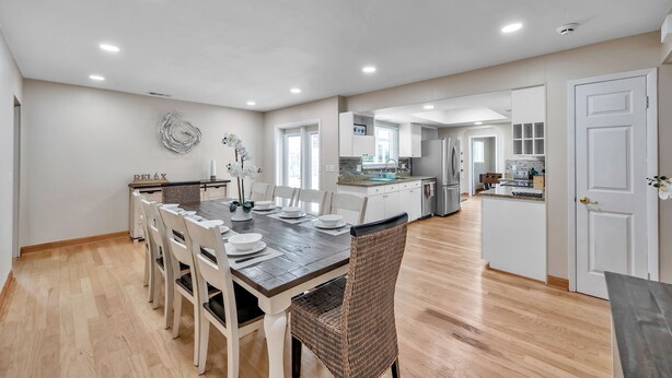 Un amplio comedor con una mesa de madera y sillas blancas, una cocina al fondo y una puerta blanca a la derecha.