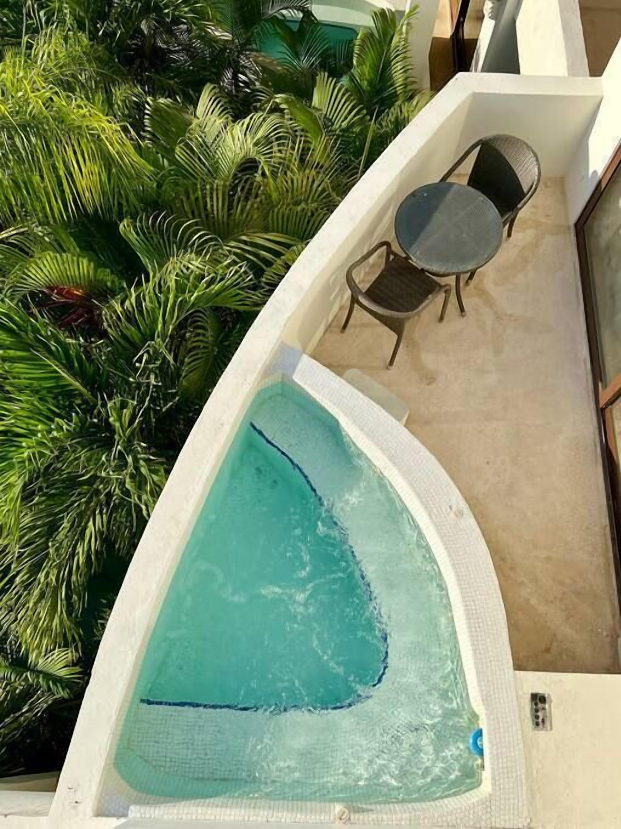 Una piscina de borde curvo, rodeada de abundante vegetación verde. Cerca, una pequeña mesa redonda y dos sillas.