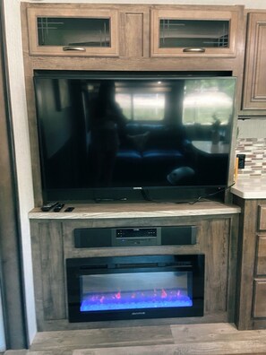 A flat-screen TV mounted on a wooden cabinet, a fireplace below it, and a kitchen area to the right.