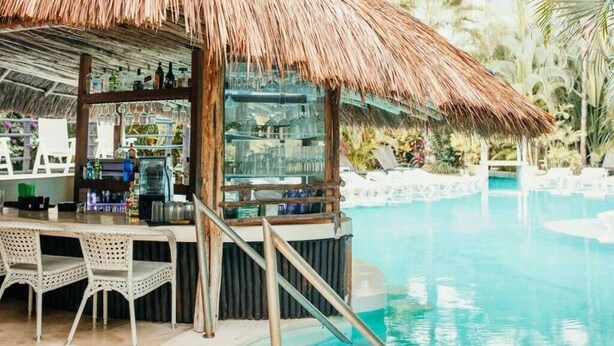 Un bar a la piscina amb teulada de palla, tamborets i una estructura de vidre.