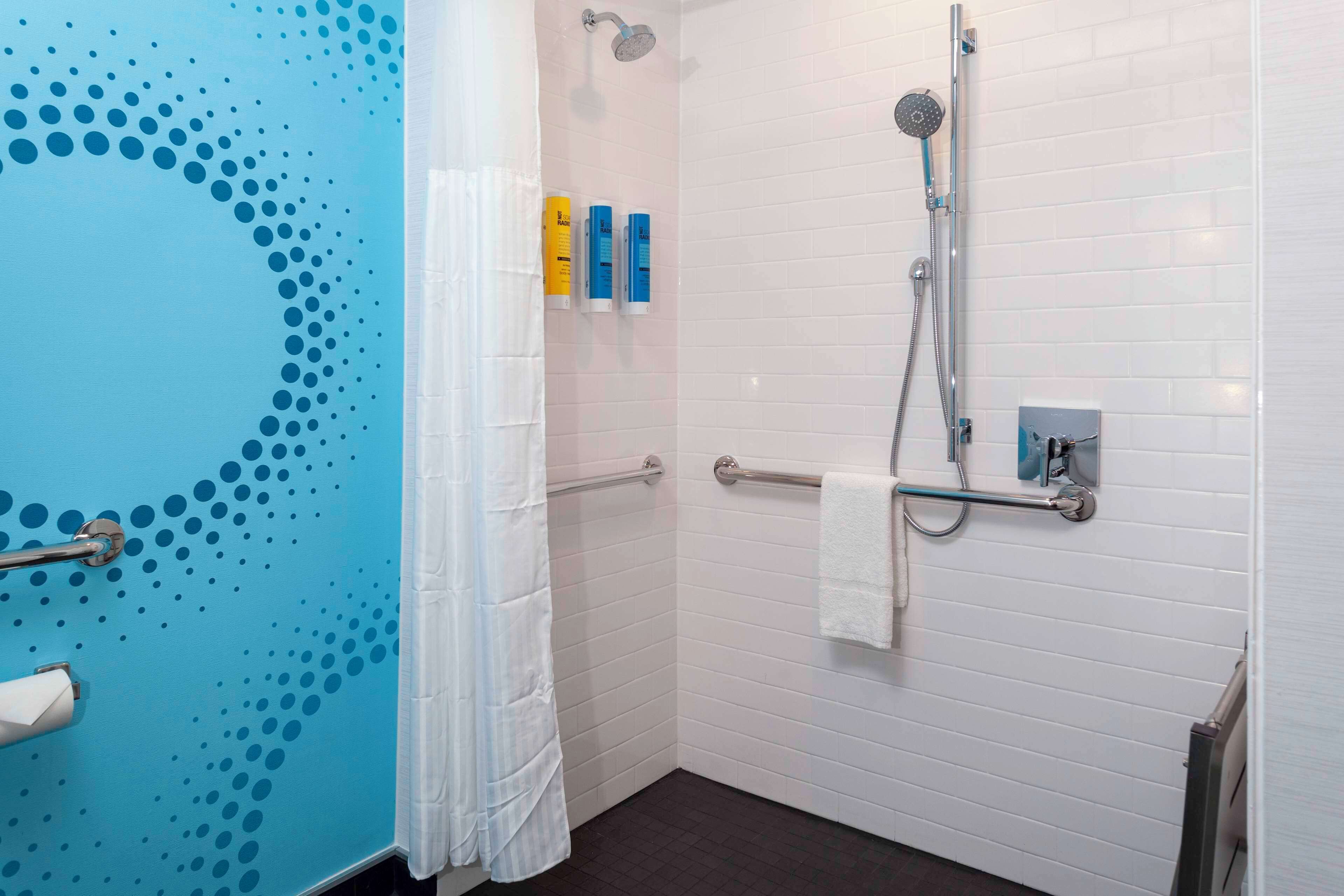 Baño con pared azul, piso de baldosas blancas, ducha con cabezal de ducha manual, toallero y dos botellas de champú.