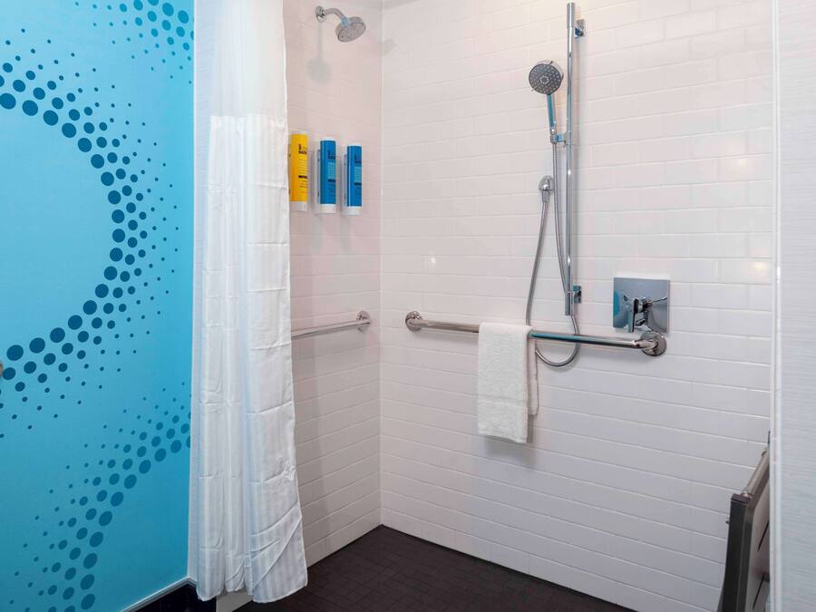 Baño con pared azul, piso de baldosas blancas, ducha con cabezal de ducha manual, toallero y dos botellas de champú.