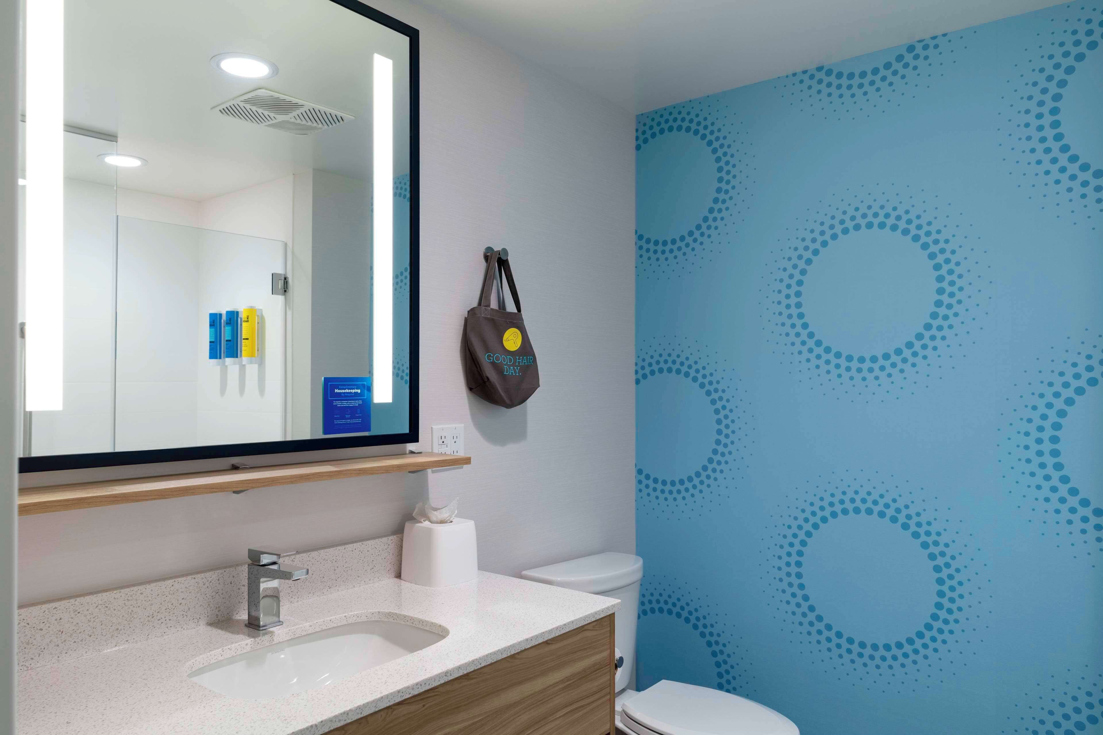 A bathroom with a white sink, mirror, and toilet. There is a towel dispenser and a bag hanging on the wall.
