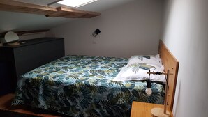 Ein kleines Zimmer mit einem Einzelbett, einem Holzkopfteil, einer Bettdecke mit tropischem Blattmuster, einem Nachttisch mit Lampe und einer kleinen Kommode.