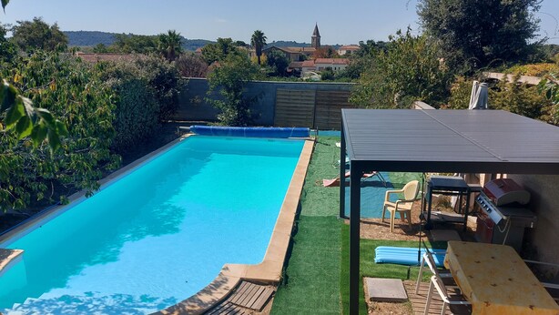 Ein Garten mit Swimmingpool, einer überdachten Terrasse und Blick auf eine entfernte Kirche.