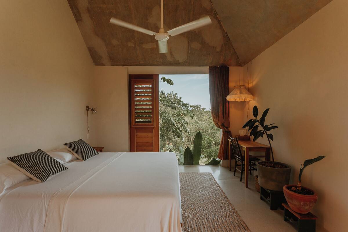 Un dormitorio con una cama, un ventilador de techo, un escritorio con una silla y una planta en maceta.