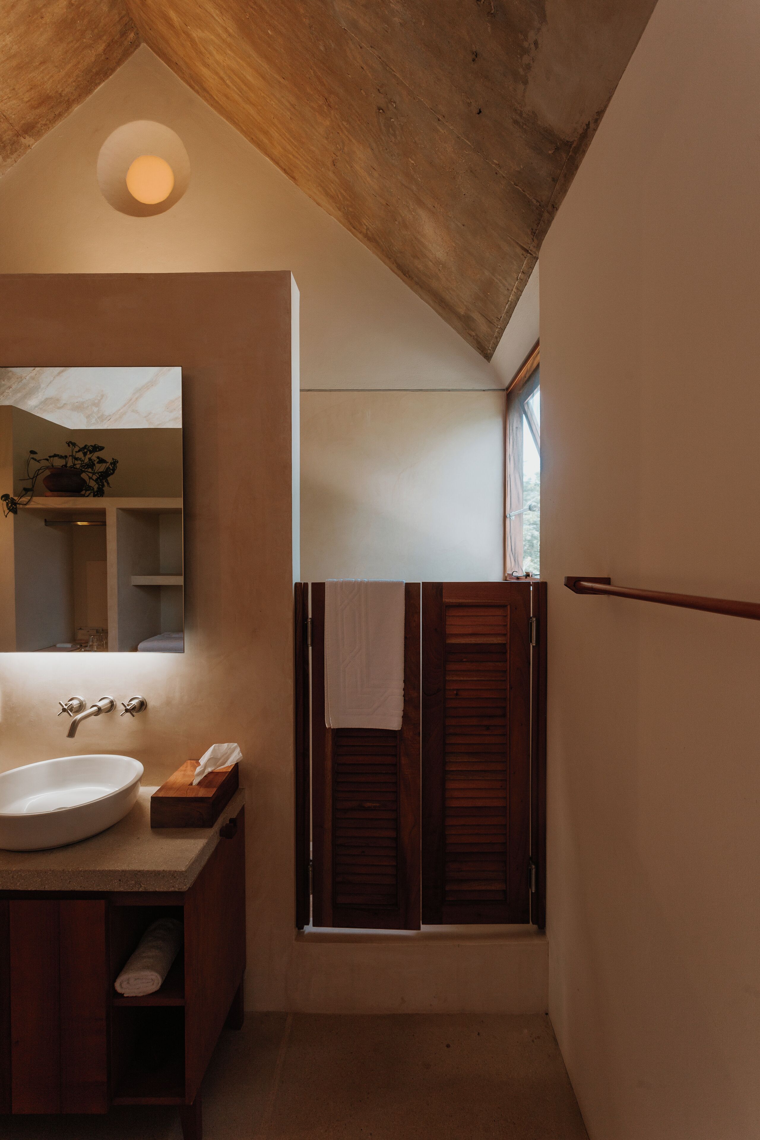 Een badkamer met een houten plafond, een grote spiegel, een wastafel en een raam met houten luiken.