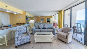 A living room with a sofa, armchair, and coffee table, a kitchen area, and a balcony with a view.