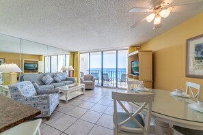 A spacious living room with a large window overlooking the sea, a ceiling fan, and a dining area with a glass table.