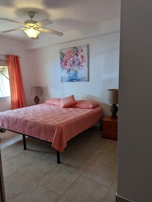 A bedroom with a bed, a ceiling fan, a painting of flowers, and a window with curtains. - Beachfront Bliss (Nueva Gorgona)