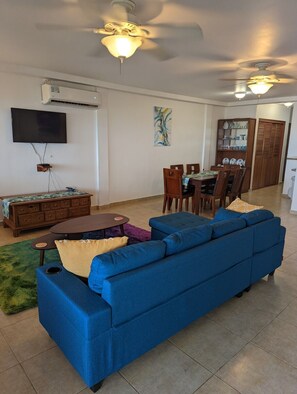 A living room with a blue sofa, a dining table with chairs, a ceiling fan, and a TV mounted on the wall. - Beachfront Bliss (Nueva Gorgona)