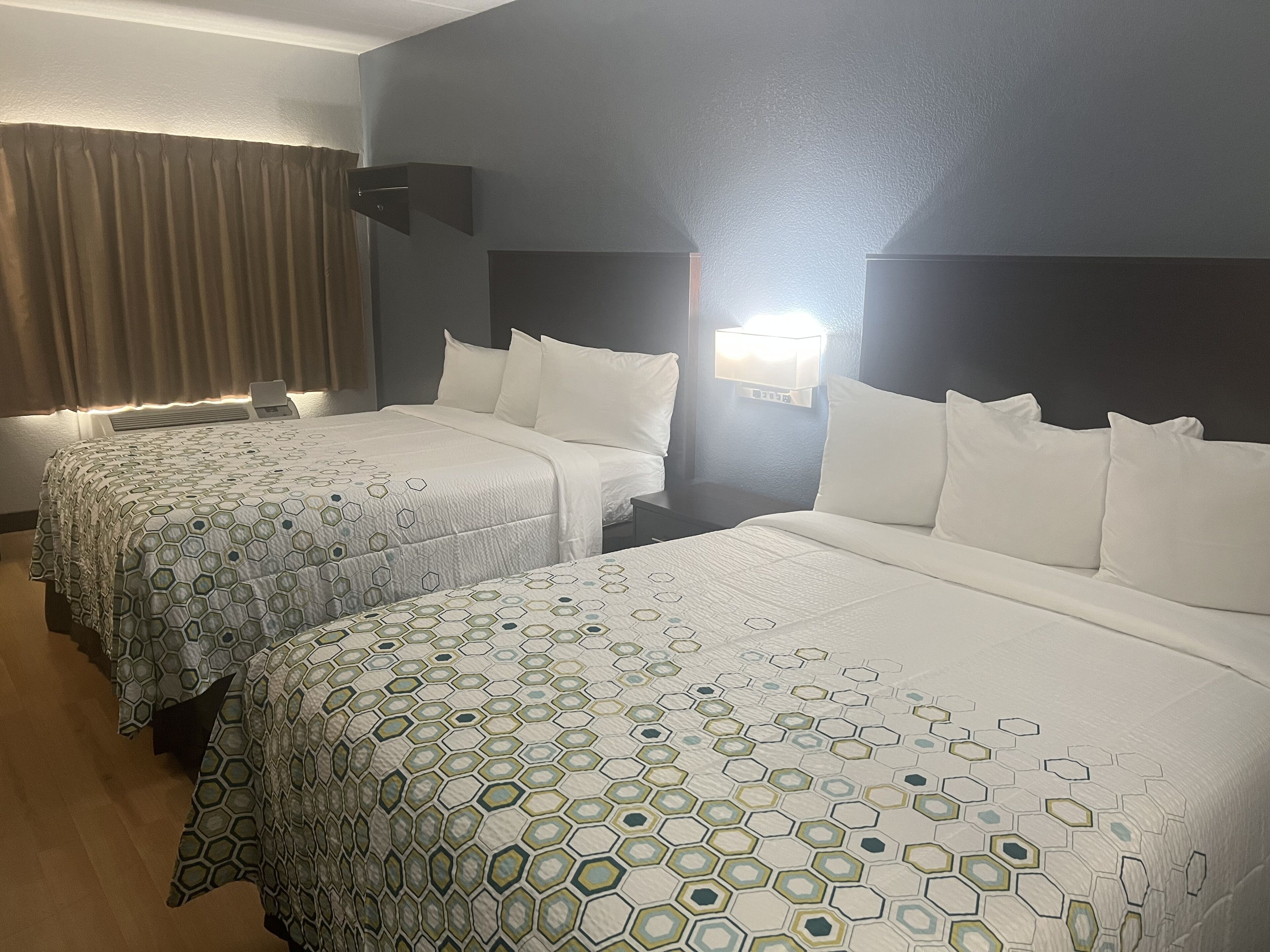 Quarto de hotel com duas camas, cada uma com colcha estampada, uma mesa de cabeceira, luminária na parede e janela com cortinas.