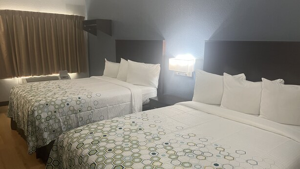 Una habitación de hotel con dos camas, cada una con una colcha estampada, una mesita de noche, una lámpara de pared y una ventana con cortinas.