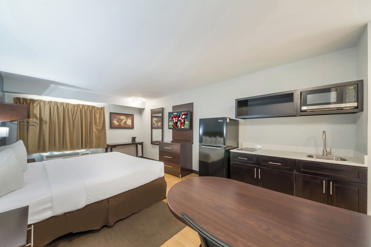 Ein Hotelzimmer mit einem Bett, einem Holztisch, einer Küchenzeile inklusive Mikrowelle, Kühlschrank und Spüle.