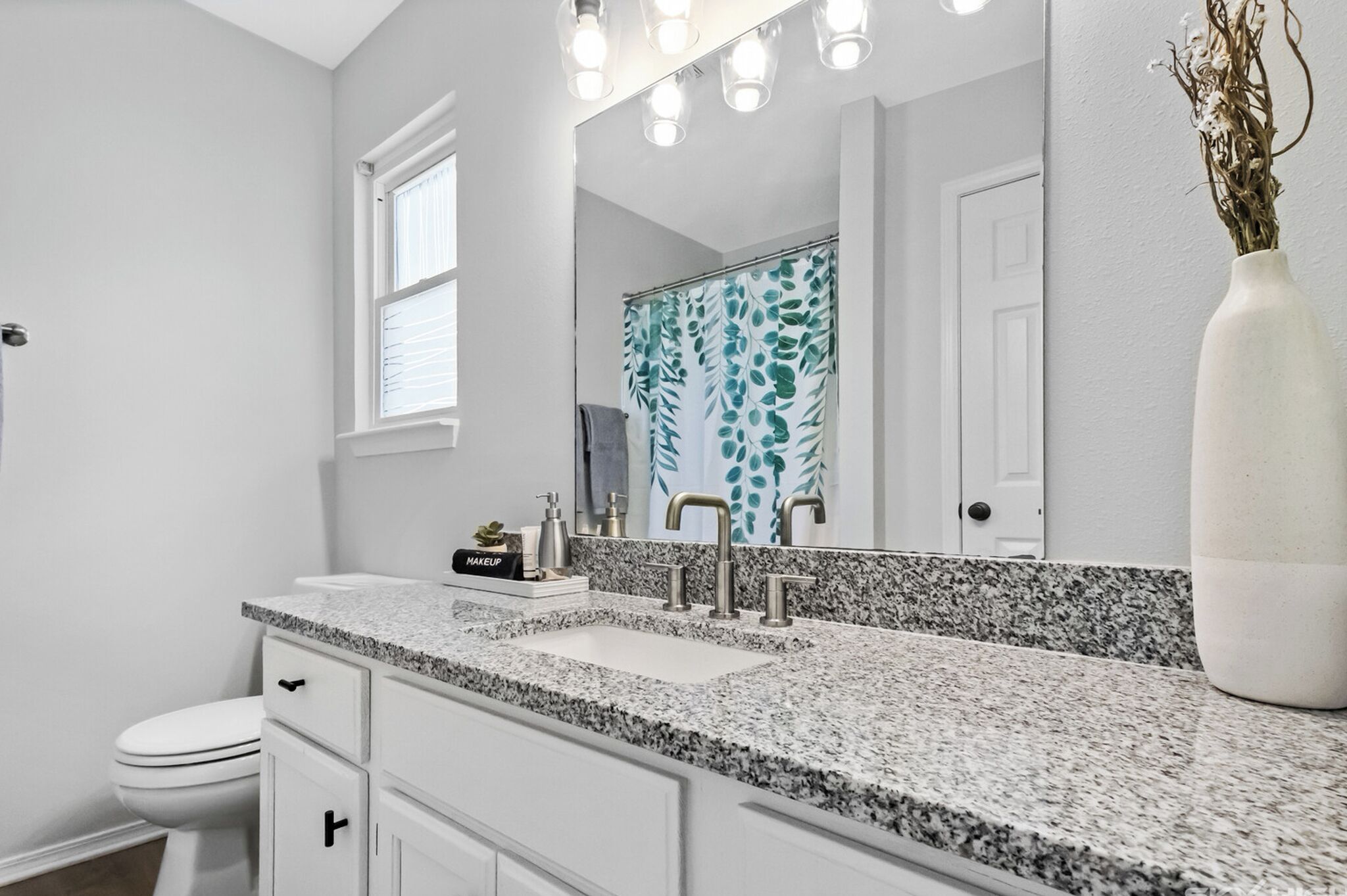 A bathroom with a granite countertop, a white sink, a mirror, and a vase with dried flowers.