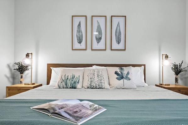 Ein ordentlich bezogenes Bett mit einem türkisfarbenen Überwurf, drei gerahmte Blätter an der Wand und zwei Nachttische mit Lampen und Vasen.