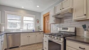 A kitchen with white cabinets, a stainless steel dishwasher, a gas stove, and a tiled backsplash. - Craftsman 4 Bed/2 Ba. Home Sleeps 10, AC & Parking (Long Beach)