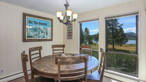 Un coin repas avec une table et des chaises en bois, un lustre, une carte accrochée au mur et une grande fenêtre donnant sur des arbres et l’eau.