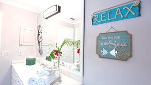 Um banheiro com toucador branco, espelho, porta-toalhas e um aviso com a frase "RELAX Two less fish in the Sea".