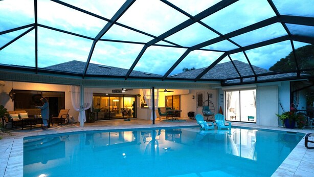 Piscina interior com teto de vidro, área de estar com cadeiras e uma mesa, e vista para o exterior.