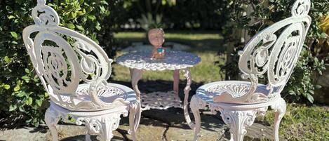 Two ornate white metal chairs and a small round table with a decorative top, set on a stone patio surrounded by greenery.