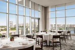 A dining area with round tables set for four, large windows offering a city view, and a carpeted floor.